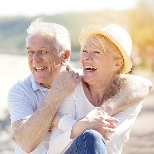 Man with white hair hugging woman in straw hat laughing on the shoreline